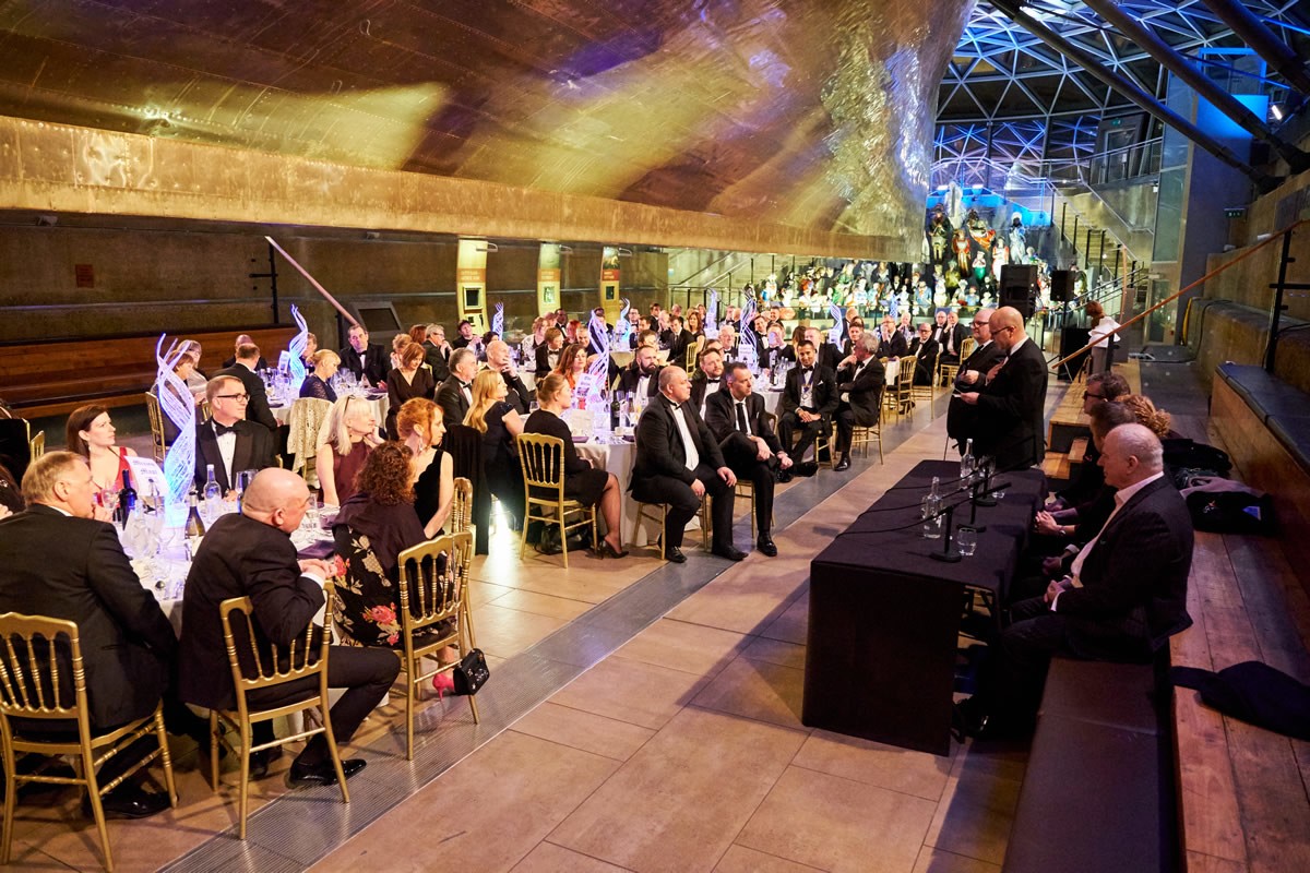a wide shot of the guests under the hull of the Cutty Sark listening to a panel of speakers